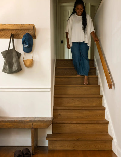 Woman walking up a wooden staircase in a home setting with bags hanging on a rack.
