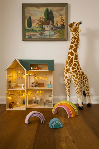 Children's toys including a dollhouse, stuffed giraffe, and rainbow toys on a wooden floor with a painting on the wall.